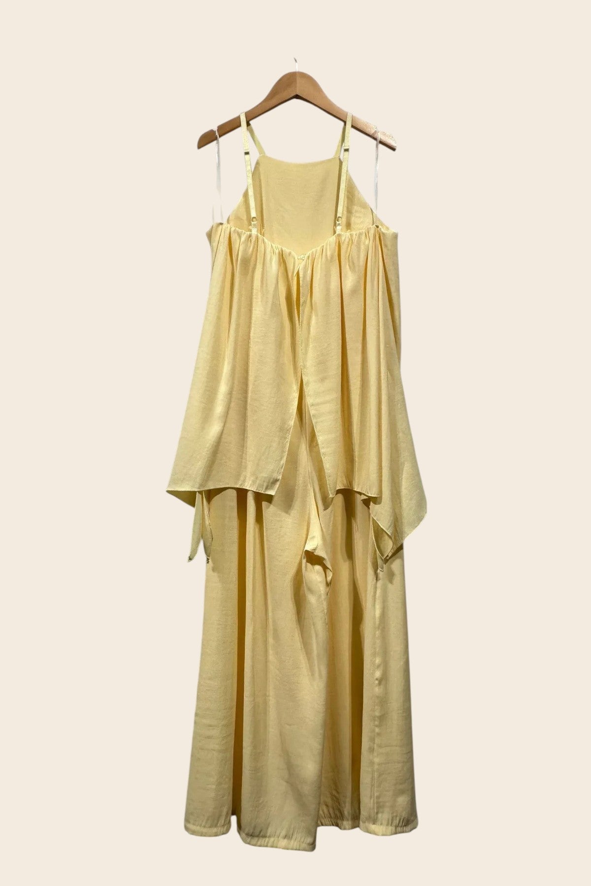 Une combinaison jaune pastel fluide et sans manches, vue de dos, suspendue à un cintre en bois. Elle présente une encolure haute et dos nu avec des bretelles réglables et des rubans transparents. Un tissu froncé crée un haut fluide et superposé, s'ouvrant sur des pantalons amples et larges qui descendent jusqu'au sol.