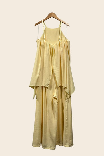Une combinaison jaune pastel fluide et sans manches, vue de dos, suspendue à un cintre en bois. Elle présente une encolure haute et dos nu avec des bretelles réglables et des rubans transparents. Un tissu froncé crée un haut fluide et superposé, s'ouvrant sur des pantalons amples et larges qui descendent jusqu'au sol.