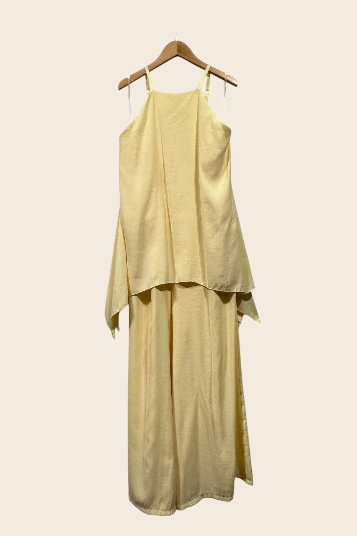 Cette image présente une combinaison pantalon pour femme, de couleur jaune pastel, vue de face. Elle est suspendue sur un cintre en bois, se détachant sur un fond uni de couleur crème clair. Le vêtement est fait d'un tissu fluide, d'aspect soyeux, avec une texture discrète.

Le haut du corps a une coupe "encolure américaine" (type halter), avec des bords supérieurs droits qui remontent haut sur la poitrine. Il est maintenu par de très fines bretelles spaghettis de la même couleur. Ce haut est composé d'un l